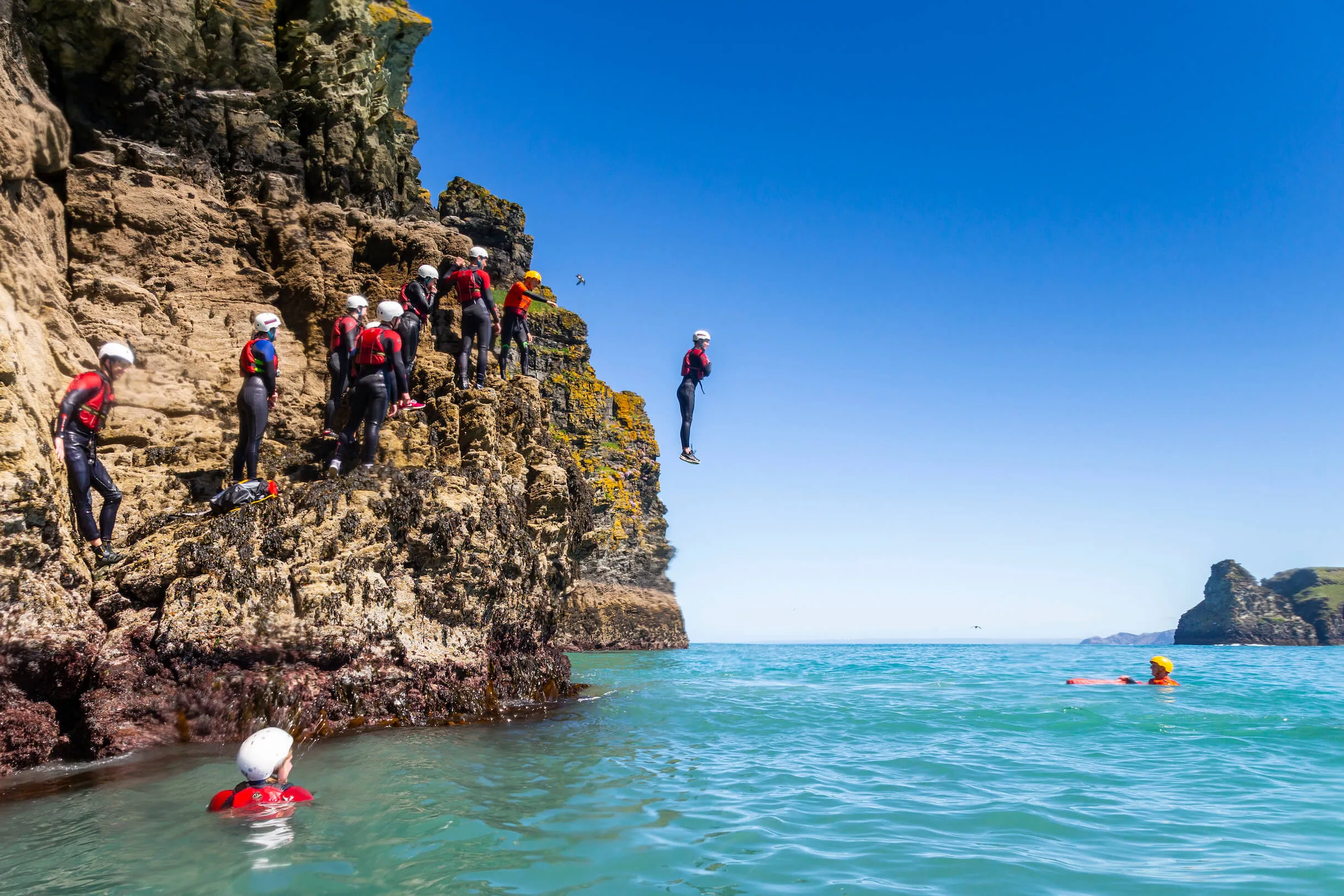 a group of people climbing and jumping safely off rocks while coasteering with two coasteer guides watching
