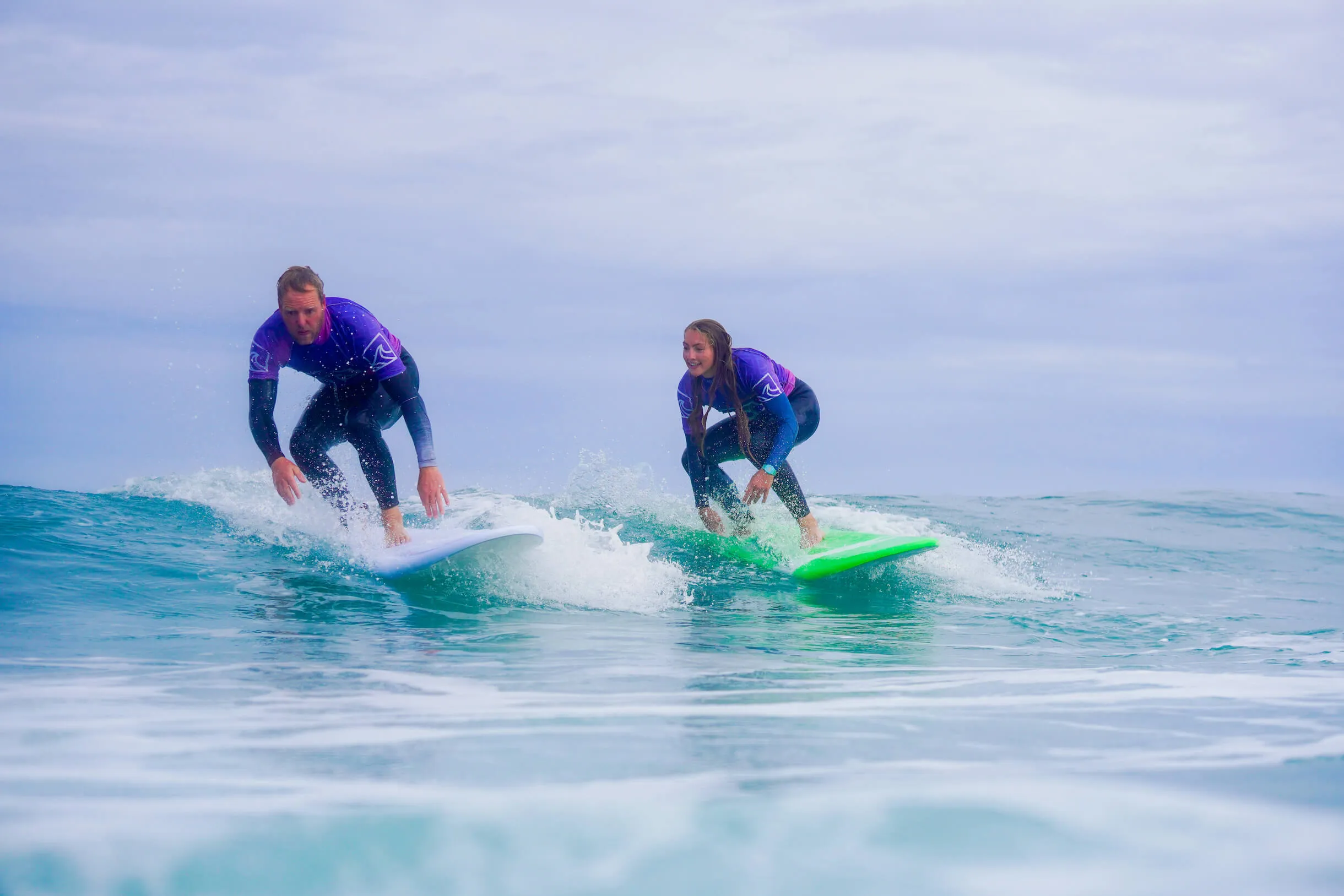 two people learning to stand up on a green wave on a surf lesson at widemouth bay